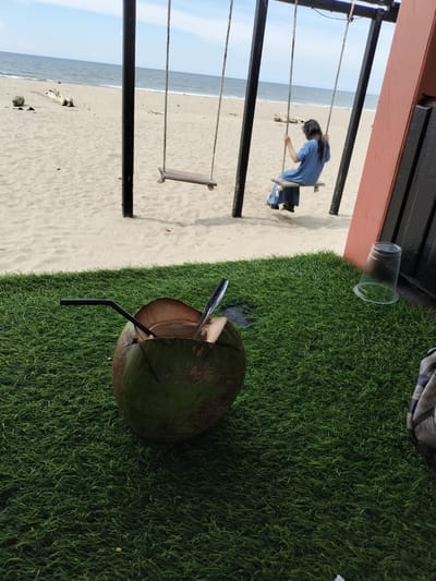 A person wearing a blue dress sits on a swing on a sandy beach with the ocean in the background. In the foreground, a halved coconut with a straw and a spoon sits on a patch of artificial grass. A clear plastic cup is also visible on the grass. The scene appears to be in Keudekruenggeukueh, Indonesia, given the coastal setting and the presence of tropical elements.