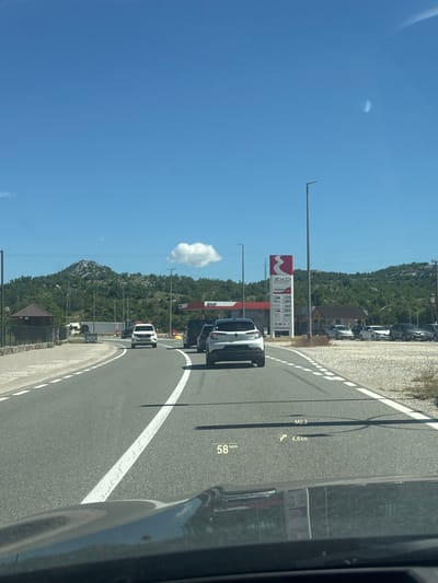 The image captures a view from a vehicle's dashboard on a sunny day, showing a road leading towards a gas station with the EKO brand. The road is asphalt, marked with white lines. Several cars are visible on the road, including a white SUV in the foreground and a white sedan further ahead. The gas station features a tall red and white sign displaying fuel prices. To the left of the road, there's a small brown building with a stone base and a wooden roof, possibly a kiosk or rest stop. In the background, there are green, rocky hills under a clear blue sky with a few scattered clouds. A head-up display (HUD) on the vehicle's windshield shows the current speed as 58 km/h, a distance of 4.6 km, and an indicator for M2.3. The image appears to be taken on the highway near Očinići, Montenegro, given the context of the EKO gas station and the surrounding landscape.