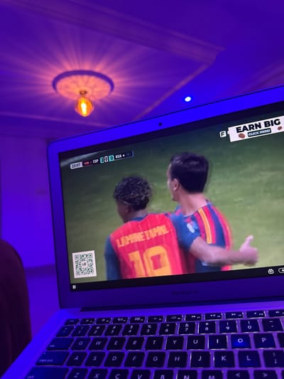 The image shows a laptop screen displaying a soccer game with the score "ESP 2-0 KSA". Two male soccer players are visible on the screen, one wearing a red jersey with the name "LAMINETAPA" and the number "10", and the other in a blue and red striped jersey. A QR code is also displayed on the screen. The laptop is positioned on a surface, and part of a keyboard is visible. The scene is illuminated by purple lighting, casting shadows and creating a dimly lit atmosphere. The lighting originates from a decorative ceiling fixture with a visible light bulb. The overall environment appears to be an indoor setting, possibly a living room or entertainment area. There is no information in the image to suggest a specific location within Laminga, Nigeria.