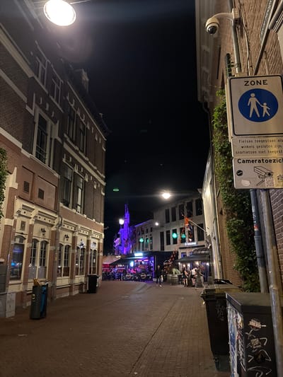 The image depicts a street scene at night in the city of Arnhem, Netherlands. On the left side of the street is a multi-story brick building with large windows and decorative white trim. Several streetlights are visible, including one prominently in the upper left corner. In the foreground, there are two trash receptacles.
On the right side of the street, a brick building wall is visible, mounted with a security camera and several signs. One sign indicates a "ZONE" with a pedestrian symbol. Another sign beneath it reads "Fietsen toegestaan als winkels gesloten zijn; snorfietsen niet toegestaan" (Bicycles allowed when shops are closed; mopeds not allowed) and "Cameratoezicht" (Camera surveillance) with a graphic of a security camera. A lamppost with a camera is also present.
Further down the street, multiple establishments with glowing lights and outdoor seating are visible. Several people are walking or standing outside these venues. Flags, including one with orange and blue colors, are displayed on some buildings, suggesting a festive atmosphere or national celebration. The street is paved with brick.