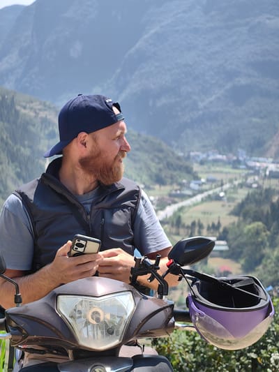 A man with a reddish beard and wearing a blue baseball cap and a dark vest stands in front of a moped in Phố Bảng Commune, Vietnam. He is holding a cell phone. A lavender helmet is hanging from the moped's handlebars. In the background are mountains and a valley with a road.
