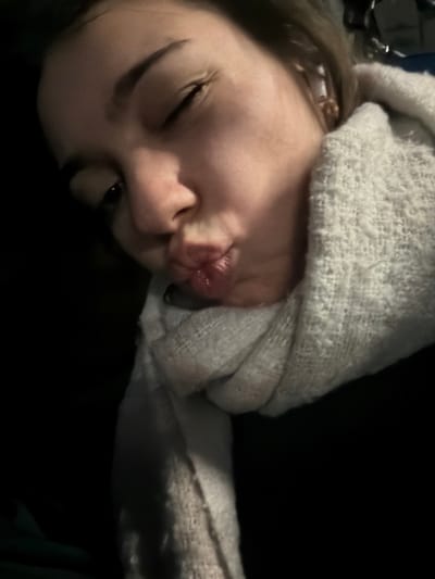 A selfie shows a person lying down with a white, textured fabric draped loosely over their head. The person's face is slightly angled, with one eye open and the other squinting. The lips are puckered, giving a whimsical expression. The background is dark and out of focus, with only hints of objects visible.