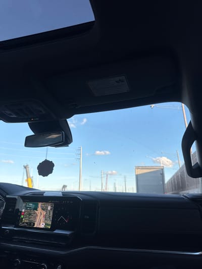 The image shows the interior of a vehicle on a sunny day. The view is from the driver's seat, looking forward through the windshield. A sunroof is visible at the top of the frame, and a rearview mirror with an air freshener hanging from it is centered above the dashboard. The dashboard features a large infotainment screen displaying a GPS navigation map and a digital clock. The screen shows a route in Elmhurst, United States of America, with the destination being a park or recreational area. Outside the vehicle, the sky is blue with scattered clouds, and there are power lines and cranes visible in the distance. A large truck is in front of the vehicle, and a fence runs along the right side of the road.