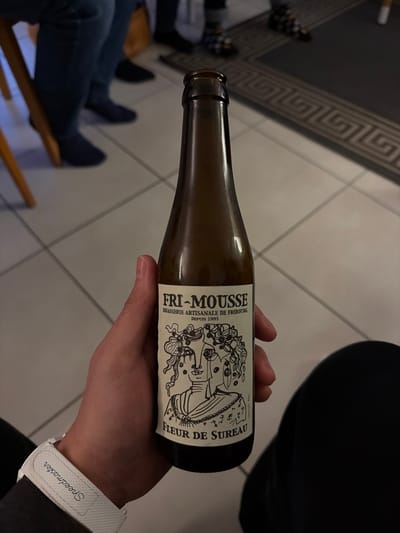 A human hand holds an uncapped, brown glass bottle of "FRI-MOUSSE" beer, with the label "BRASSERIE ARTISANALE DE FRIBOURG DEPUIS 1993" and "FLEUR DE SUREAU" visible, adorned with a stylized black and white illustration of a face. A white wristband with "Speedmaster" text is visible on the wrist. The background depicts an indoor setting with light-colored tiled flooring and dark grout lines. Partially visible are the lower legs and feet of several individuals, including one person wearing dark blue trousers and blue socks/shoes, and another wearing patterned socks and dark shoes. A section of a light-colored wooden chair leg is also visible. A dark rug with a light Greek key border pattern is present on the floor in the upper right background. The scene is located indoors in Givisiez, Switzerland.