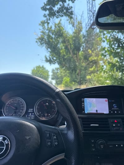 The image shows the interior of a car, likely a BMW, as indicated by the steering wheel emblem. The driver's perspective is captured, with the steering wheel prominently in the foreground. The dashboard displays include a speedometer, tachometer, and a navigation screen showing a map. The car appears to be driving on a road surrounded by lush green trees and vegetation, suggesting a rural or suburban setting, possibly on a sunny day given the bright sky. The overall scene suggests a drive through a scenic route, potentially within or near the city of Belene, Bulgaria, as the context of the prompt specifies. There are no people visible in the image, and the car is the central object of focus.
