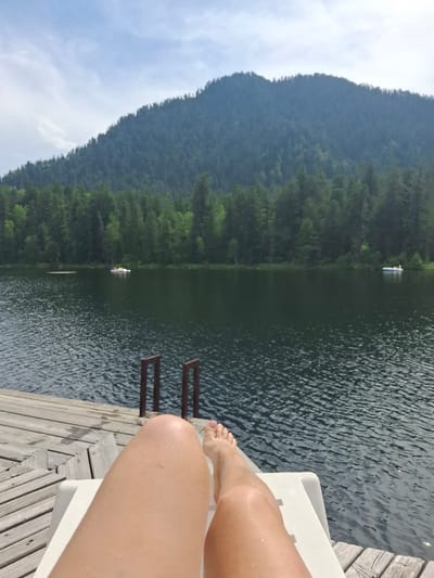 A person's legs are in the foreground, resting on a white lounge chair on a wooden dock. The dock extends into a dark, rippling lake. In the background, a dense, dark green forest covers the slopes of a large mountain under a partly cloudy sky. There are two small, white watercraft visible on the lake in the middle ground. The scene appears to be in Vydrino, Russia.