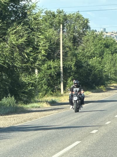 In the city of Дорожный, Russia, two individuals are seen riding a motorcycle on a paved road. The person in the front is wearing a black helmet and a black t-shirt with a graphic design. They are operating the motorcycle. The second person is seated behind the driver. The motorcycle is traveling on a road with a gravel shoulder on the left side. Lush green trees line the side of the road, and in the background, buildings and a water tower are visible on a hill under a clear sky. The scene is illuminated by daylight, casting shadows on the road surface.