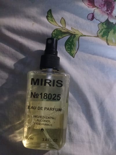 A clear glass bottle of "MIRIS N°18025 EAU DE PARFUM" is pictured from a slightly high angle. The bottle contains a pale yellow liquid and has a black spray nozzle. The label on the bottle also lists "INGREDIENTS: ALCOHOL FRAGRANCE", and "100 ml 3.4 FL. OZ". The bottle rests on a white fabric with a floral pattern. The background is dimly lit and shows a pattern of light and shadow. This scene is not located in the city of Hlushky, Ukraine.