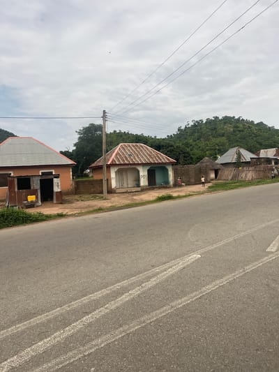 The image shows a roadside scene in Minda, Nigeria. A paved road with white markings runs diagonally across the right side of the frame. On the left, a line of buildings with corrugated metal roofs sits alongside the road, backed by a densely forested hill. One building, painted light blue and white, has the word "POS" visible on its facade. Another structure appears to be made of stacked concrete blocks with a thatched roof, and a person in a light-colored shirt stands near it. A partially visible building in the foreground has a metal roof and what looks like a small kiosk attached. Power lines stretch across the cloudy sky. The lighting suggests it is daytime, possibly overcast, and the presence of vegetation indicates a warm climate.