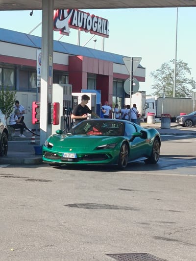 A green Ferrari sports car with a red interior is parked at a gas station with "AUTOGRILL" signage. A young man in a black t-shirt is standing next to the driver's side of the car, holding a smartphone and appearing to interact with the vehicle. Two people are seated on a bench to the left of the gas pumps. Several other individuals are standing and walking near the Autogrill building in the background. The scene appears to be outdoors on a sunny day, with a paved road and gas pumps visible. The license plate on the Ferrari reads "HB-571SZ." The image was taken in Tortona, Italy.