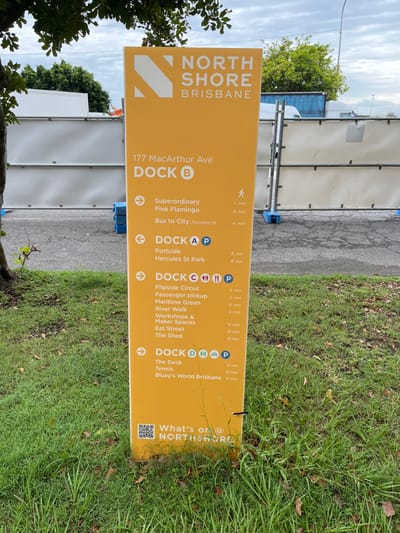 A large, upright, yellow wayfinding sign for "NORTH SHORE BRISBANE" stands outdoors in what appears to be a mixed-use or developing area in Hamilton, Australia. The sign features destinations, estimated walking times, and directional arrows.

The top of the sign displays "NORTH SHORE BRISBANE" and "177 MacArthur Ave". Below this, it lists various locations categorized by "DOCK" with associated walking times and icons.

**DOCK B** points to:
*   Superordinary (1 min)
*   Pink Flamingo (6 min)
*   Bus to City Theodore St (4 min)

**DOCK A** (with a parking icon) points to:
*   Portside (8 min)
*   Hercules St Park (8 min)

**DOCK C** (with parking, accessible toilet, and possibly dining icons) points to:
*   Flipside Circus (5 min)
*   Passenger pickup (5 min)
*   Maritime Green (6 min)
*   River Walk (6 min)
*   Workshops & Maker Spaces (6 min)
*   Eat Street (8 min)
*   The Shed (10 min)

**DOCK D** (with parking, accessible toilet, and possibly dining icons) points to:
*   The Deck (time unreadable from OCR, but visible as 11 min on the image)
*   Tennis (12 min)
*   Bluey's World Brisbane (12 min)

The bottom right corner of the sign shows "What's on @ NORTHSHORE" with a QR code.

The setting includes a paved surface in front of the sign, leading to a grassy area on the right. In the background, there are temporary construction barriers or fences, and some green foliage from trees. The sky is overcast, suggesting daytime. The sign's purpose is to guide pedestrians to various points of interest within the North Shore Brisbane precinct.