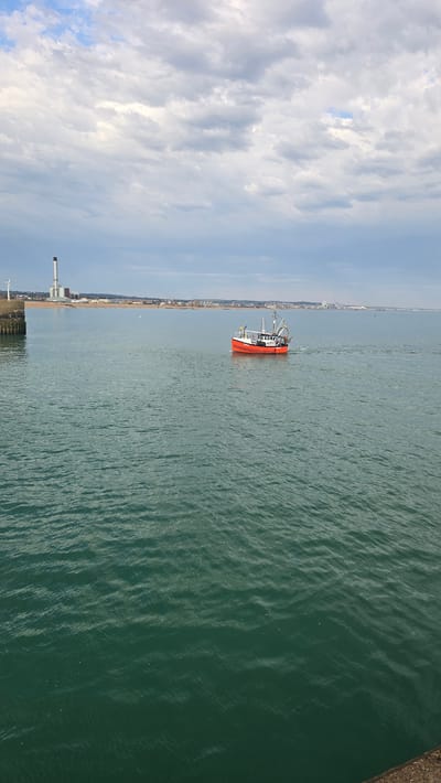 A fishing boat, predominantly red with a white superstructure, travels across a body of water under a cloudy sky. The boat is oriented diagonally, moving from left to right in the frame, leaving a slight wake. In the background, a distant shoreline is visible, featuring buildings and a tall industrial structure with a chimney. To the left of the frame, a section of a stone pier juts out into the water, with wooden pilings visible at its edge. The water itself appears choppy with a dark green hue. The scene is set in Shoreham-by-Sea, within the Adur district of the United Kingdom.
