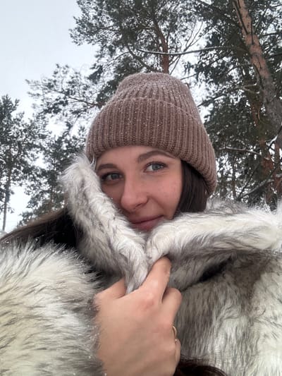 An outdoor portrait captured in a wooded area of Kalinovo, Russia, depicts a female subject from the upper chest upward in winter conditions. The subject is a young adult female with dark hair, visible below a light brown ribbed knit beanie speckled with white particles, likely snow or glitter. She has light skin, blue eyes, and a slight smile. She is wearing a thick, two-tone faux fur coat with shades of white, grey, and black. Her right hand is visible, adorned with a gold-colored ring on the ring finger, pulling the fur collar of her coat upward to cover part of her chin and mouth. Small white particles, consistent with snow, are present on her beanie and the fur of her coat. The background consists of tall pine trees with snow-dusted branches against an overcast, light grey sky.