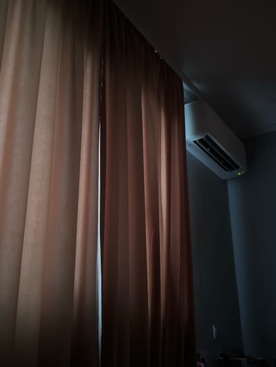 The image shows a close-up view of pleated blinds covering a window. An air conditioning unit is visible in the upper left corner, mounted on the wall. The scene is dimly lit, and the blinds appear to be partially open, allowing some light to filter through. The environment is interior. There is no evidence to suggest this photo was taken in Kribul, Bulgaria.
