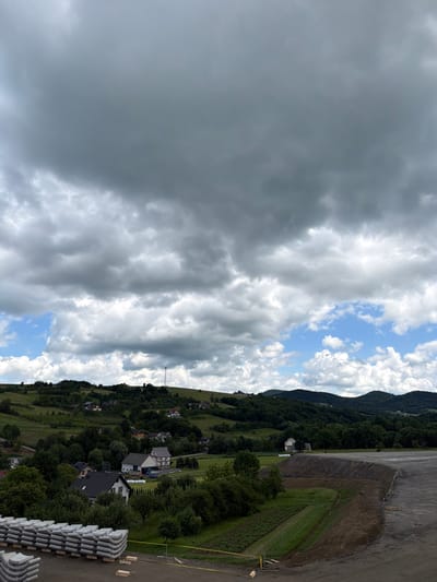 This is a photograph taken on a cloudy day in Jodłownik, Poland. The image shows a rural landscape with rolling green hills, scattered houses, and a large area of prepared ground in the foreground. In the foreground, a stack of white, curved concrete elements is visible on a dirt surface. Further back, a partially constructed embankment is covered with dark gravel and yellow caution tape. Beyond this, the landscape transitions to lush green fields, interspersed with trees and residential buildings. A small village or hamlet is nestled in the valley, with houses having red or gray roofs. The background features a series of forested hills under a dramatic sky filled with dark and light gray clouds, with patches of blue sky visible. A slender telecommunications tower stands on one of the distant hills. The overall impression is one of rural development or construction in a scenic, hilly terrain.