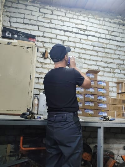 A man wearing a black t-shirt and pants, and a black baseball cap, stands with his back to the camera, drinking from a large plastic bottle of water. He is in what appears to be a workshop or storage area. The background is a white brick wall. Shelves are visible, stacked with cardboard boxes, some with labels in Cyrillic script. To the left, there is a tall, beige metal cabinet. On a workbench in front of the man, there are a pair of gloves and a pack of Marlboro cigarettes. The floor is cluttered with tools and equipment, including what looks like an orange-colored piece of machinery, possibly part of a generator or engine. The lighting suggests an indoor, possibly dimly lit, environment, with a light source visible on the ceiling. The scene is consistent with a storage area or workshop in a city like Shimanovsk, Russia, given the presence of Cyrillic labeling.
