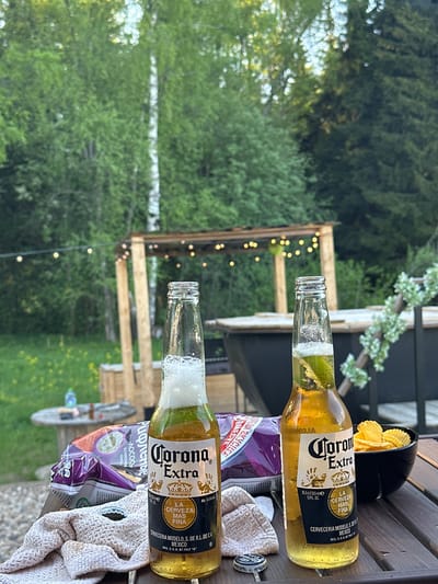 This image shows an outdoor setting, possibly a patio or deck, with two bottles of Corona Extra beer prominently displayed. The beers are accompanied by a bowl of chips and a bag of potato chips, suggesting a casual gathering. The background features lush greenery, including trees and foliage. There is no indication that this image was taken in Малеевка, Russia.