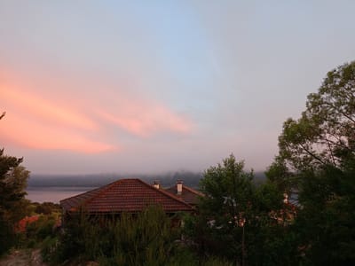 An outdoor, low-angle, wide shot captures two tiled-roof houses on a hillside overlooking a body of water, likely Lake Sarnitsa, Bulgaria, during a hazy sunset. The sky is a gradient of soft pinks, oranges, and blues, with wispy clouds tinged with the sunset colors. The foreground is dominated by dense green foliage and the roofs of the houses. The house on the left has a red tile roof and is partially obscured by trees. The house on the right is larger, with a prominent red tile roof and two chimneys emitting faint white smoke. Beyond the houses, a misty landscape of rolling hills meets the calm water of the lake. The overall atmosphere is serene and slightly melancholic due to the fading light and haze. There are no people visible in the image.