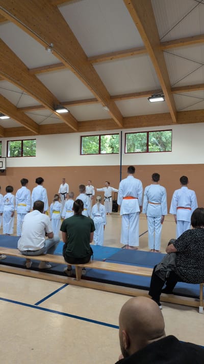 A group of people are standing in a gymnasium in Neudietendorf, Germany, facing a man in a white karate uniform with an orange belt, who is gesturing with his hands. Several children and adults, also in white karate uniforms, are lined up. Some have yellow belts, while others have no belts visible. A few adults are seated on a wooden bench in the foreground, facing away from the camera. The gymnasium has a wooden ceiling with exposed beams and lighting fixtures. Windows with trees visible outside are present on the far wall. The floor is a light color with blue lines.