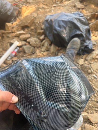 The image shows a close-up of a person's hand holding a black, carbon-fiber-patterned object, likely a welding mask. The mask has a silver checkered pattern and what appears to be the letters "MSG" imprinted on its surface in a lighter color. The background is out of focus, revealing a rocky, outdoor environment with debris, including a black plastic bag and a plastic bottle. A person's boot is visible in the upper right quadrant, suggesting the person holding the mask is wearing work boots. The image appears to be taken in Kresna, Bulgaria, given the rocky terrain and vegetation typical of the region.