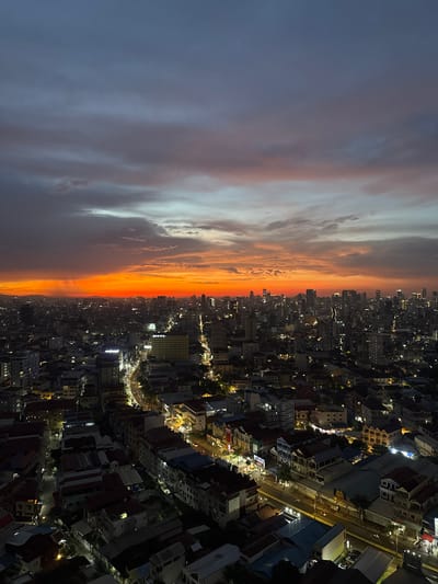 The image depicts a panoramic view of Khan Chamkar Mon, Cambodia, at dusk. The sky is a dramatic canvas of color, transitioning from a deep indigo at the top to a fiery orange and red horizon. Streaks of light, possibly sun rays, pierce through the lower clouds. Below the vibrant sky, the cityscape unfolds, a dense urban landscape illuminated by the twilight. Numerous buildings of varying heights, from low-rise structures with red roofs to taller modern towers, are visible. Streetlights and building lights create a network of glowing points across the city, indicating a developed urban environment. Roads are visible with illuminated streaks suggesting vehicular traffic. The overall scene conveys the transition from day to night in a bustling city.