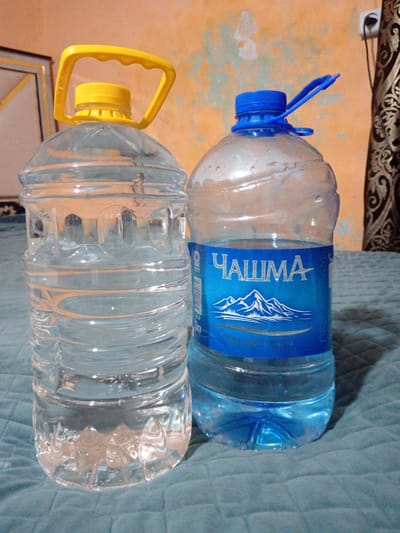 Two large plastic bottles of water are placed on a quilted teal-colored surface. The bottle on the left has a yellow cap and handle. The bottle on the right has a blue cap and a blue label featuring the word "ЧАШМА" (Chashma) in white Cyrillic letters, with an illustration of mountains. The background is an orange-colored wall with some lighter patches. The scene is indoors, and based on the provided location, it is in the city of Заргар, Tajikistan.