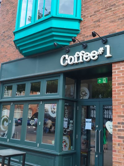 This image shows the exterior of a "Coffee #1" establishment in Wychavon, United Kingdom. The building is constructed of red brick, with a prominent teal-colored bay window on the upper level. Below the bay window, a dark green facade houses the shop's entrance and windows. White lettering on the dark green facade spells out "Coffee #1". Two black outdoor lights are mounted above the lettering. The windows display reflections of the street, including people and vehicles. One window features a green emergency exit sign with running figures and arrows. A heart-shaped sign with the words "See You Soon" is visible on the right side of the entrance. A small table with a dark top and metal legs is positioned to the left of the entrance.