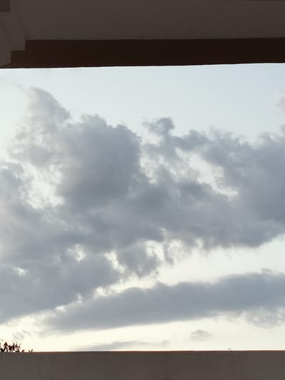 This media file shows a cloudy sky as seen from beneath a building overhang. The sky is a mix of light gray and white clouds, with some areas of brighter white suggesting sunlight breaking through. The overall impression is of an overcast or partly cloudy day. In the foreground, the edge of a brown overhang is visible, casting a shadow on the top portion of the frame. Below the sky, a flat, tan-colored surface, likely a wall or roof, stretches across the bottom of the image. On the far left of this surface, silhouetted against the sky, are several small, dark figures. These figures appear to be small plants or possibly decorative objects. To the right of these figures, there are no other discernible objects on the surface. The lighting suggests it is either early morning or late afternoon, given the soft light and the presence of clouds. The scene appears to be an outdoor setting, possibly a balcony, rooftop, or courtyard, with a view of the sky.
