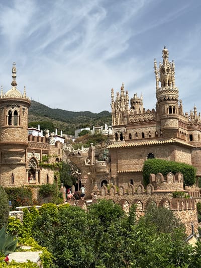 The image depicts a stone castle-like structure built on a hillside with lush green vegetation in the foreground. The castle features intricate architectural details, including turrets, arches, and decorative stonework. Several people are visible on staircases and pathways within the castle complex. In the background, houses and a green, mountainous landscape are present under a partly cloudy sky. This structure is identified as Castillo de Colomares, a monument located in Benalmádena, Spain, dedicated to Christopher Columbus and the discovery of America.