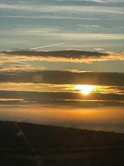 Golden sunlight pierces through cloudy formations in an ethereal, vertical composition. Sky is blue and a wispy contrail cuts diagonally across the left side. To the right, shadows are cast by dark objects out of focus. The atmosphere is filled with a sense of warmth.