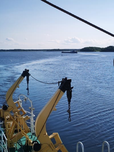 The image depicts a view from a vessel sailing on a calm, blue body of water, likely a river or lake, under a clear sky with scattered white clouds. In the foreground, yellow cranes with black components are visible, suggesting the vessel is equipped for loading or unloading operations. The cranes are positioned on the deck of the ship, which has a green painted hull. In the distance, a long barge or cargo ship is being towed, leaving a visible wake. The shoreline is lined with lush green trees. The scene is likely set in Nova, Russia, given the context provided, possibly on the Volga River, a major waterway in Russia. The water appears to be relatively undisturbed, with small ripples on the surface. A dark, thick cable traverses the top of the frame, originating from the upper right and extending towards the left, suggesting it is part of the ship's rigging. No people are clearly visible in the image.