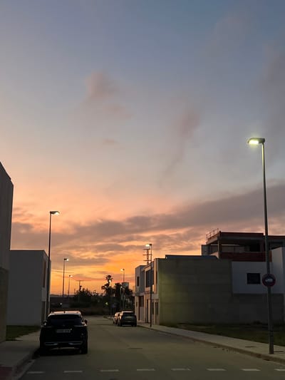 This image depicts a residential street in the city of Alcàsser, Spain, at sunset or dusk. The sky transitions from a pale blue at the zenith to vibrant orange and pink hues near the horizon, with scattered clouds illuminated by the setting sun. Multiple active streetlights, emitting bright yellow-white light, punctuate the scene.

On the left side of the street, a dark-colored SUV, bearing the Spanish license plate "3990 MVM," is parked parallel to the curb, facing away from the viewer. Further down the street on the right, two additional dark-colored vehicles are parked. The street itself is paved, with white dashed lines marking sections of the road.

Modern, minimalist residential buildings with flat roofs line both sides of the street. Structures are primarily white and grey, with some exhibiting exposed concrete or reddish-brown brick on upper levels, suggesting ongoing construction or recent development. A tall, white building is partially visible on the far left. On the right, several buildings are present, one featuring an upper-level balcony railing and another displaying a circular red and white "no parking" road sign.

In the middle distance, silhouetted against the bright horizon, are trees, including palm trees, and utility poles with visible lines. Sidewalks, composed of light-colored paving stones, flank the street. No people are visible in the frame.