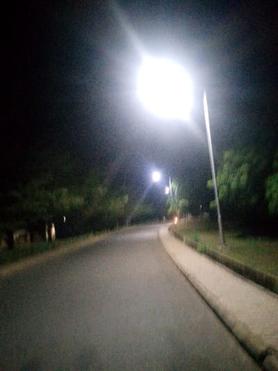 A night scene depicts a curving road lit by streetlights. The closest light is intensely bright, creating a halo effect. The road surface appears smooth, flanked by a raised concrete curb on the right. Lush greenery borders both sides of the road. Further down, additional streetlights provide softer illumination along the bend. The overall atmosphere is quiet and dimly lit, with the bright lights serving as beacons.