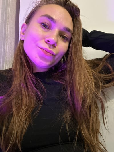A medium shot depicts a woman with light skin and brown hair. Her face is illuminated with a purplish-pink light. She wears a black shirt or jacket. The background is a neutral white. The location is not discernible from the image.