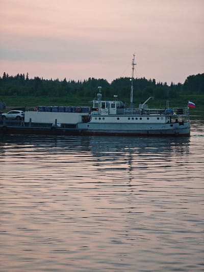 This image captures a wide shot of a ferryboat on a calm body of water, likely a river, at dusk. The sky is painted with soft, pale pink and light grey hues, indicating either sunrise or sunset. A dense line of dark green trees forms the backdrop on the distant shore, suggesting a natural, possibly remote, setting. The water's surface is rippled, reflecting the subdued colors of the sky and the silhouette of the boat.

The ferry itself is a substantial vessel, primarily light grey with a darker red hull visible at the waterline. It appears to be transporting cargo, with what looks like several rows of large, rectangular packages stacked on its deck. A white car is also visible on the deck, positioned towards the front. The ferry has a superstructure with a mast, windows, and railings. A Russian flag is prominently displayed at the stern of the boat.

Text is visible on the side of the ferry's superstructure, partially obscured but readable as "ECT-794". The overall mood of the image is peaceful and serene, characterized by the quiet water and the soft evening light. The scene suggests a routine ferry operation, possibly in a rural or natural landscape like Krivlyak, Russia, as indicated by the location context. The weather appears to be clear and calm, contributing to the tranquil atmosphere.