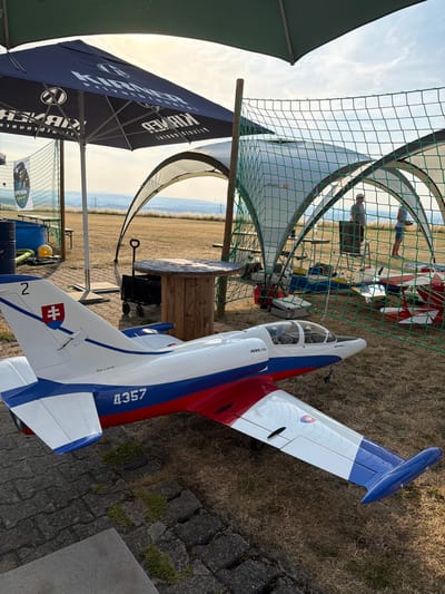 An outdoor scene depicts a model airplane on a paved area in front of a grassy field. The model airplane is white, blue, and red, with the number "4357" on its fuselage and a Slovak flag on its tail. In the background, under a green canopy and netting structure, several other model airplanes are visible, as well as two individuals standing. A large black umbrella with white lettering is to the left. The overall setting suggests an event or gathering related to model aviation. The location is identified as Becherbach, Germany.