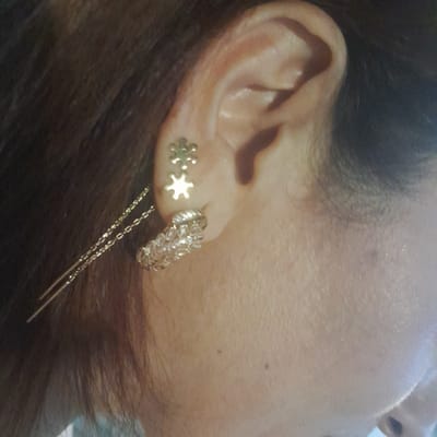 A close-up profile shot shows a person's ear with multiple earrings and hairpins. Two small, silver, snowflake-shaped stud earrings are visible on the upper part of the earlobe. Below these, a larger, circular, textured silver hoop earring is worn. Two thin, silver hairpins with chain-like detailing are inserted into the hair behind the ear. The person has dark brown hair. The background is out of focus, showing the texture of skin and hair. The lighting is soft, with a slight sheen on the skin. This image is not representative of any specific location in Rudəkənar, Azerbaijan.