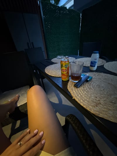 The image depicts a dimly lit outdoor patio area in Nea Peramos, Greece, likely at dusk or night. A person's legs and hand are in the foreground, with the hand wearing a ring and resting on their lap. In the background, a black table is set with three woven placemats. On the table are a can of Lipton Peach iced tea, a glass with a dark amber liquid, an e-cigarette device, a pack of cigarettes, and a bottle of white liquid with blue labeling. Behind the table, dark chairs are visible, and the patio is enclosed by a green hedge and dark wooden fencing. The overall impression is one of a relaxed evening setting.