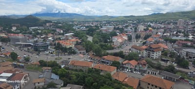 This is an aerial view of a city, with a prominent river flowing through it. The city is nestled in a valley with rolling green hills and mountains in the background, partially obscured by clouds. The architecture features a mix of older, smaller buildings with terracotta roofs and some modern, multi-story structures. The streets are lined with trees and appear to be moderately busy with cars. A gas station with a prominent canopy is visible, as is a modern building with a glass facade. The landscape suggests a temperate climate. The image appears to be taken from a high vantage point, possibly a drone or a tall building, looking down on the urban sprawl and surrounding natural environment.
