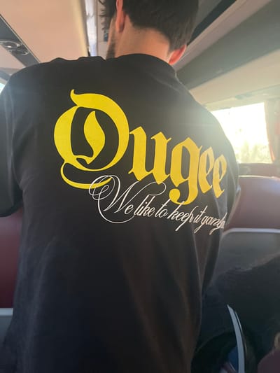 The image depicts the back of an adult male seated within what appears to be a public transport vehicle, such as a bus or train, in Leiria, Portugal. The male has short, dark hair with a fade haircut and visible stubble or a short beard on his neck. He is wearing a black long-sleeved t-shirt with text printed on the back. The prominent text, in a large yellow gothic-style font, reads "Ougee." Below this, a secondary line of text in white cursive font is partially visible, reading "We like to keep it gar..." The interior environment includes light-colored overhead panels or ceiling, and dark red or maroon seatbacks are visible on either side of the subject. Bright sunlight enters through windows on the right side of the frame, creating a lens flare on the t-shirt. A portion of another passenger's dark clothing is visible in the lower right corner. The individual is oriented away from the camera, and no specific actions or interactions are discernible.