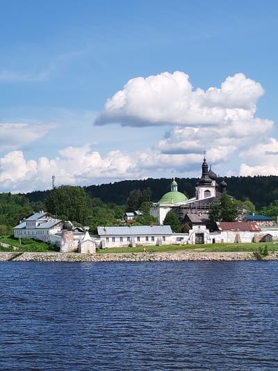 The image shows a tranquil scene of the Goritsky Monastery on the banks of the Volga River near Goritsy, Russia. In the foreground is the rippled surface of the dark blue river. A low rocky shoreline separates the water from the monastery buildings. The monastery is a collection of primarily white buildings with various architectural styles, including green and black domes topped with Orthodox crosses. Scaffolding is visible on one of the church structures, indicating ongoing restoration work. The monastery buildings are framed by a backdrop of lush green trees, providing a contrast to the white structures. A transmission tower is also visible amidst the trees. Above the monastery and trees, a blue sky is punctuated by large, fluffy white clouds.