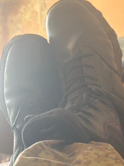 The image shows a close-up of two black leather boots, with one boot placed on top of the other. A camouflage-patterned garment is visible to the right of the boots. The laces of the top boot are visible. The image is taken with low lighting and unclear focus. There is no information that indicates this image was taken in Dolna Mitropolia, Bulgaria.