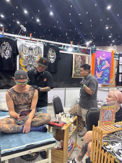 The scene is an indoor tattoo booth at a convention in the city of Ban Rong Ruea Kham, Thailand. Four individuals are present. In the foreground-left, a male client, heavily tattooed across his torso and legs, sits cross-legged on a light blue examination table, wearing a grey cap, reflective green sunglasses, and camouflage shorts with a blue compression wrap on his left leg. He holds a small, dark object and looks down, smiling slightly. Behind him and to his right, a male tattoo artist, wearing a black cap, black t-shirt, denim shorts, and black gloves, applies a tattoo to the client's right shoulder with a machine, smiling at the client. To the right, a second male, also tattooed, stands wearing a grey t-shirt, plaid shorts, and black gloves, with an ID badge, looking rightward. Further right, a female with pink hair and a small facial tattoo is seated at a table, observing. A clear acrylic stand displaying a QR code for "@@NHATBE" is positioned on the table in front of her. A two-shelf wooden utility cart holds tattoo supplies, including bottles, liquids, and a red box, situated between the client and the seated female. The booth's rear wall displays black t-shirts featuring designs such as a stylized face, intertwined cobras with "NHATDE" text, and a skull. Also visible are a poster with floral vase motifs and a vibrant purple banner with butterfly designs. The overhead ceiling features exposed structure and numerous track lighting fixtures.