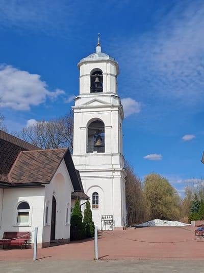 The image captures a white Orthodox church bell tower against a blue sky with scattered clouds. The tower has multiple tiers with arched openings revealing large bells. A brown tiled roof of a smaller building is visible at the base of the tower, and part of a white building adjoins it. Trees are visible in the background. A red brick-paved area and a white wall run along the right edge of the image. The location is identified as Ivashevo, Russia.