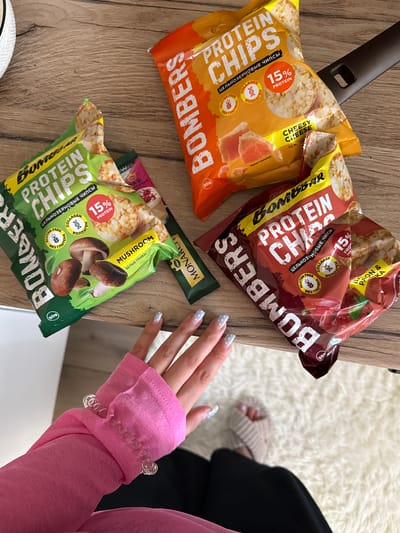 A close-up overhead shot shows several bags of "Bomber Protein Chips" arranged on a wooden surface, possibly a table, next to a fluffy white rug. The bags are primarily orange, red, and green, with prominent text in English and Russian advertising "Protein Chips" and flavors like "Cheesy Cheese," "Becons & Ricoh," and "Mushroom." Some bags indicate "15% Protein," "Gluten Free," and "Collagen."
In the foreground, a person's hand, adorned with light blue nail polish with white polka dots, is reaching towards the bags. The person is wearing a bright pink long-sleeved top with a ruffled cuff, and a clear, coiled bracelet is visible on their wrist. Part of the person's arm and shoulder are in view, along with a glimpse of their legs and feet, wearing beige socks, on the rug. The lighting suggests it's daytime. The scene appears to be indoors, possibly in a living room or kitchen area in Tliustenkhabl', Russia, given the location context. The arrangement of the snack bags and the presence of the hand suggest a casual moment of someone about to enjoy a snack.