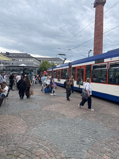This image depicts a bustling urban square in Darmstadt, Germany, identifiable by the prominent "Langer Ludwig" monument (tall reddish-brown column) in the background. The scene features numerous pedestrians, a tram, and multi-story buildings under an overcast sky.

A modern, articulated tram, primarily white with blue and orange accents, occupies the right side of the frame, appearing to be at a stop with passengers visible through its windows. Overhead electrical lines for the tram span across the sky.

On the left and in the middle ground, approximately twenty to thirty people are visible. Many are walking across the cobblestone-paved square, which features a distinct fan or arc pattern in shades of grey and reddish-brown. Several individuals are carrying bags, including a woman in the foreground with a grey tote bag and another behind her with a large blue shopping bag. Two people are seated at an outdoor cafe table on the far left, partially obscured by a yellow parasol. A person on a bicycle is also visible on the left.

In the middle ground, a glass-enclosed structure houses a Starbucks, with a "Steig 1" (Platform 1) sign indicating a public transport stop. Behind this, several functional multi-story buildings with numerous windows form the backdrop. The "Langer Ludwig" column, a tall brick structure topped with an observation platform, rises prominently on the far right, partially obscured by the tram. The sky is uniformly cloudy.