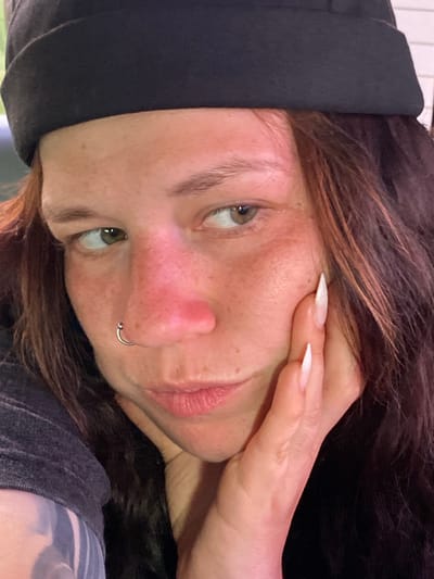 The image is a close-up, self-taken portrait of a person wearing a black beanie and a dark grey t-shirt. The individual has fair skin with freckles, prominent reddish flushing on their cheeks and nose, and long, dark brown hair. They have a silver hoop nose ring. The person is resting their right hand on their cheek, with long, white acrylic nails visible. Their gaze is directed slightly to the left. The background is dark and out of focus. There is no identifiable urban environment present in the image that would allow for a specific location in Верхнеозёрное, Russia to be determined.