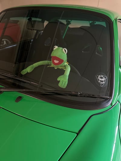 A Kermit the Frog plush toy is positioned on the dashboard of a bright green car, its arms outstretched. The car appears to be a sports car, possibly a Porsche, with black racing seats visible through the windshield. A sticker for the "Sports Car Club of Helsinki" is affixed to the lower right corner of the windshield. The scene is illuminated by artificial light, suggesting it is indoors, likely in a garage. The background is blurred, but appears to show other vehicles and storage shelving, consistent with a private garage setting in Espoo, Finland.