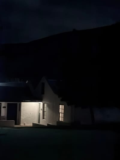 The image depicts a stark night scene, likely an urban alleyway or internal courtyard, in the Overstrand Local Municipality, South Africa. When viewed in a corrected vertical orientation, a multi-story building facade dominates the right side of the frame, rendered in pale, possibly white or light grey tones. This building is illuminated by artificial lights emanating from within, casting a warm glow through several windows featuring dark horizontal and vertical grilles. Architectural details such as ledges, recessed sections, and a possible air conditioning unit or vent are visible on its surface.
The left side of the image is enveloped in profound darkness, suggesting another unlit building or a deep shadow, which contrasts sharply with the brightly lit facade. This creates a dramatic interplay of light and shadow, emphasizing the geometric forms of the structures. No people or specific activities are discernible, contributing to a quiet, solitary atmosphere characteristic of late night. There is no visible text. The overall mood is one of urban stillness, highlighted by artificial light piercing through the pervasive darkness.