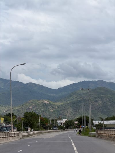 The image displays a panoramic view of a road winding alongside a lush, green mountain range under a heavily overcast sky. The scene is oriented horizontally, with the vast expanse of mountains dominating the left side and a paved road with an adjacent developed area on the right.
The sky is filled with dense, grey clouds, indicating an overcast daytime. The mountains are rugged and covered in dense vegetation, appearing verdant and imposing. Some rocky formations are visible among the trees. Towards the foreground and right side of the image, an asphalt road with clear white lane markings stretches into the distance. A white dashed line separates what appear to be two lanes, while a solid white line marks the shoulder.
A sturdy concrete guardrail runs alongside the road, separating it from the lower terrain and scattered buildings. Several streetlights with long, curved arms extend over the road, accompanied by multiple power lines supported by utility poles. Along the roadside, there are a few buildings, some partially hidden by trees, suggesting a degree of human settlement or commercial activity. A couple of motorbikes or scooters with riders are visible on the road, indicating light traffic. One motorbike is more prominent in the mid-ground.
The overall impression is one of a modern road infrastructure carved through a natural, mountainous landscape, typical of regions like Cam Ranh Ward, Vietnam, where natural beauty coexists with development. No legible text is visible on any signs.
